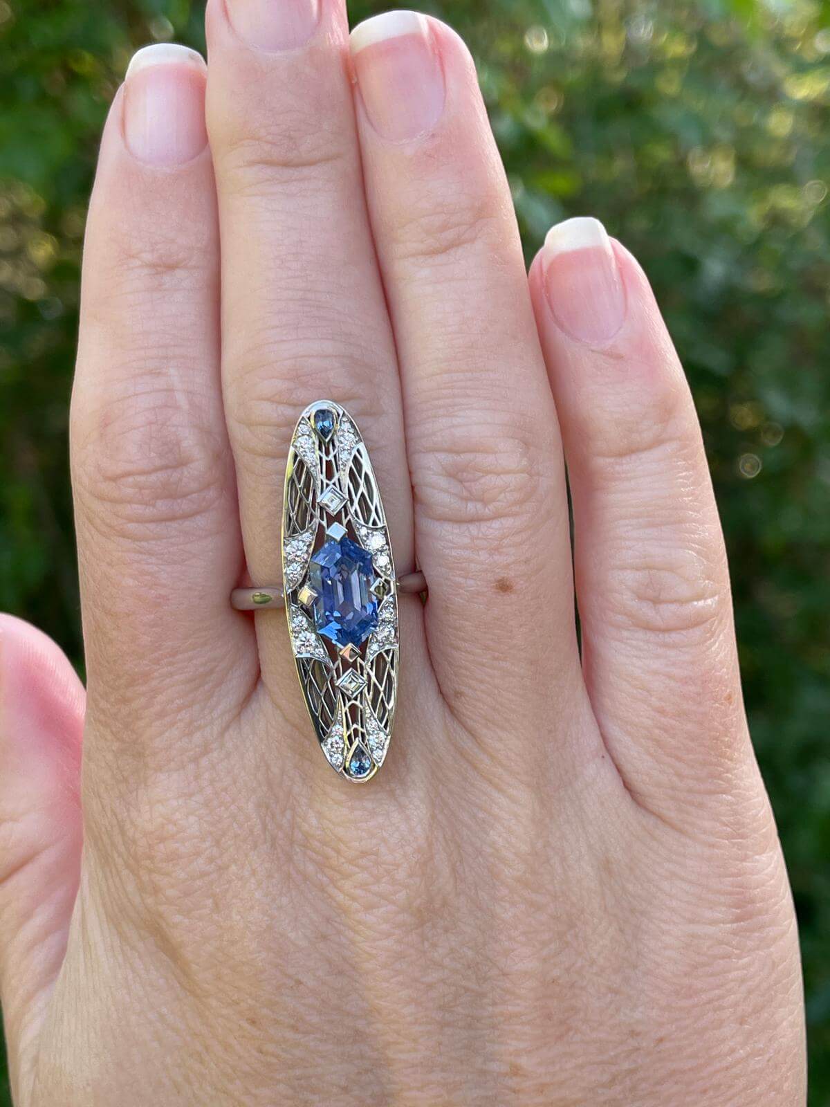 Pictured on a hand is a platinum ring featuring an unheated elongated hexagon Ceylon sapphire in a four-prong square setting, in an arrangement of carré and round diamonds. Two petite pear-shaped blue sapphires sit on either side of the centre stone. Handmade by Sydney jeweller Nicky Burles.