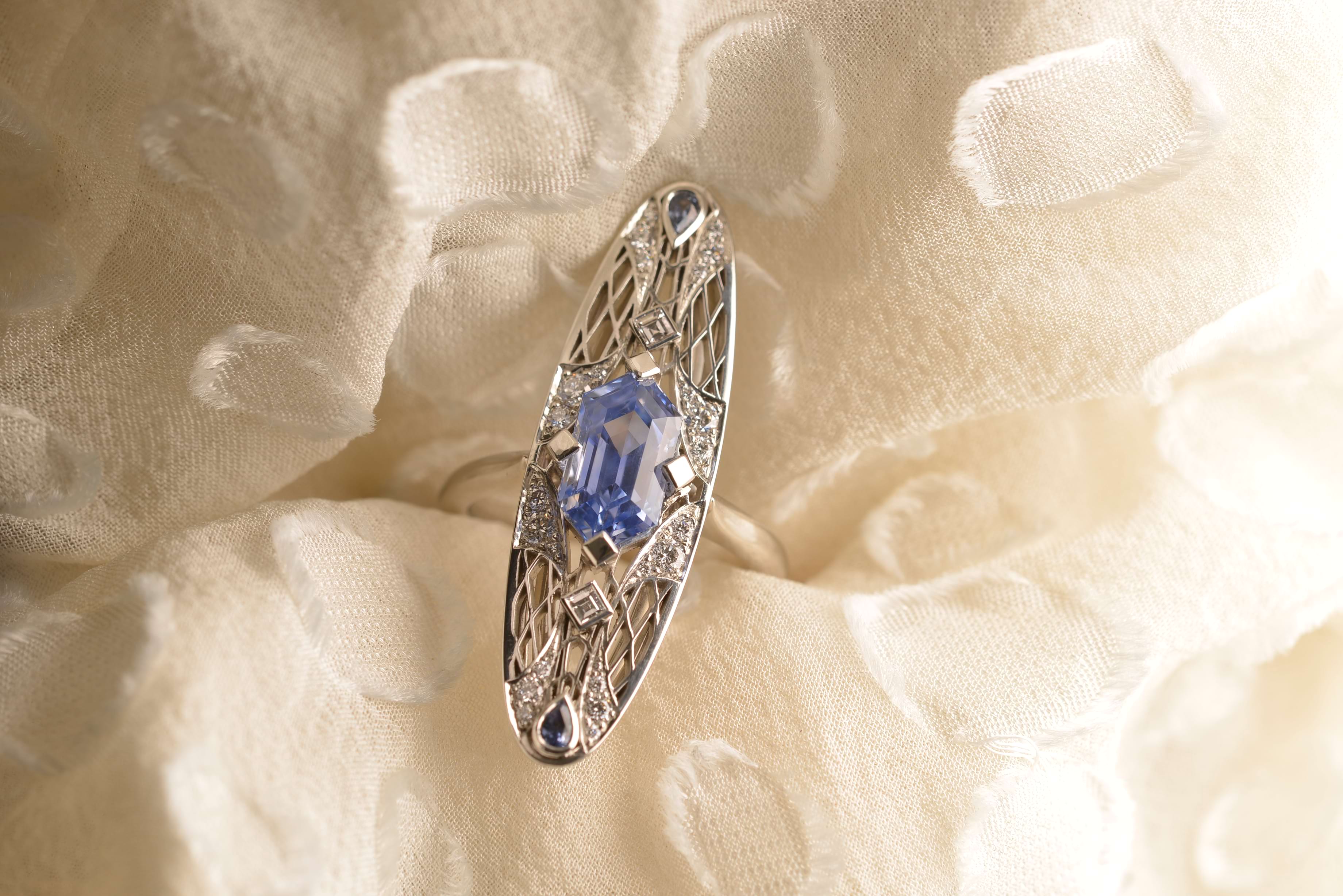 Alae on fabric background. Platinum ring featuring an unheated elongated hexagon Ceylon sapphire in a four-prong square setting, in an arrangement of carré and round diamonds. Two petite pear-shaped blue sapphires sit on either side of the centre stone. Handmade by Sydney jeweller Nicky Burles.